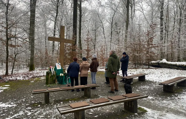 Trauernde an einer Waldbestattung im Winter: ein Holzkreuz, davor eine grün verhüllte Ablage mit Kerzen und einem Blumengesteck, ringsum Steinbänke im verschneiten Buchenwald